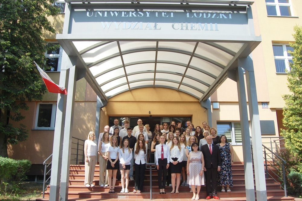 Grupa osób stojąca na schodach przed budynkiem Wydziału Chemii Uniwersytetu Łódzkiego. Uczestnicy zdjęcia są elegancko ubrani większość z nich ma na sobie białe koszule lub bluzki. Po lewej stronie widoczna jest flaga Polski. Otoczenie budynku jest zadbane, z zielenią w tle. 
