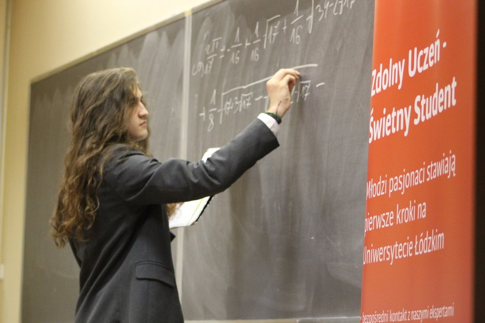 Młody mężczyzna z długimi, falowanymi włosami, który pisze kredą na dużej tablicy w sali wykładowej. Ubrany jest w ciemny garnitur, co nadaje mu elegancki wygląd. Mężczyzna skupia się na matematycznych wzorach, które zapisuje na tablicy. Obok tablicy znajduje się czerwony baner z napisem "Zdolny Uczeń - Świetny Student" oraz informacjami o programie, który wspiera młodych pasjonatów w stawianiu pierwszych kroków na Uniwersytecie Łódzkim.