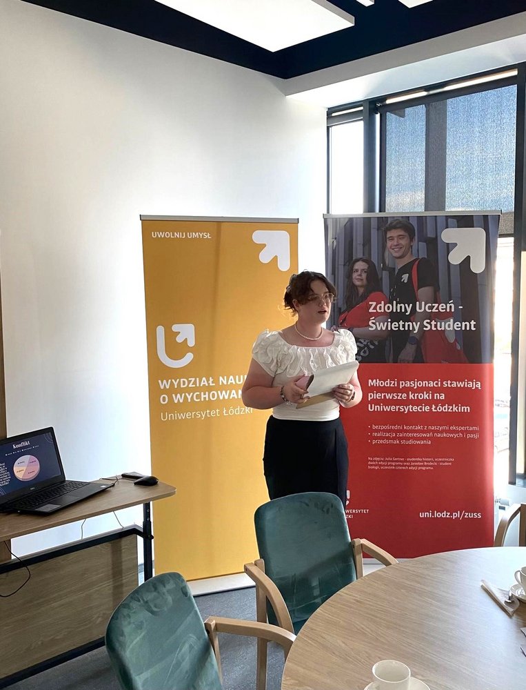 Kobieta w sali konferencyjnej, ubrana w białą bluzkę i czarną spódnicę, trzyma dokumenty w rękach i uśmiecha się do kamery. W tle widać dwa roll-upy reklamowe: : jeden żółty z logo Wydziału Nauk o Wychowaniu, drugi czerwony z logo programu Zdolny uczeń-świetny student. Obok kobiety znajduje się stolik z laptopem. W pomieszczeniu jest duże okno, przez które wpada naturalne światło.
