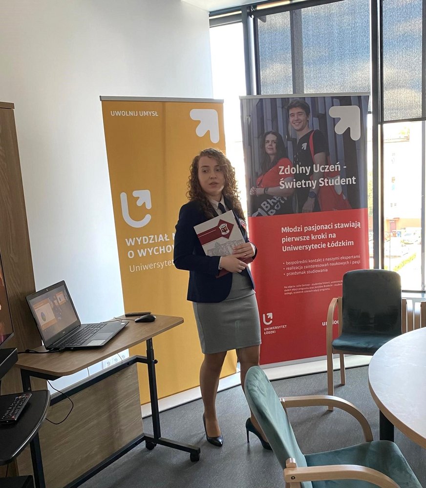 Na zdjęciu znajduje się kobieta w sali konferencyjnej. Jest ubrana w elegancką marynarkę i spódnicę, trzyma dokumenty w rękach i uśmiecha się do kamery. W tle znajdują się dwa roll-upy: jeden żółty z logo Wydziału Nauk o Wychowaniu, drugi czerwonyz logo Zdolny uczeń-świetny student. Obok kobiety znajduje się stolik z laptopem, a w tle widać duże okna, przez które wpada naturalne światło. Pomieszczenie jest jasne i nowoczesne, z krzesłami ustawionymi wokół okrągłego stołu.