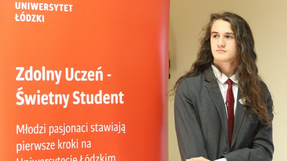 Młody mężczyzna stojący przed czerwonym roll-upem z napisem "Uniwersytet Łódzki" oraz sloganem "Zdolny Uczeń - Świetny Student". Mężczyzna ma długie, ciemne włosy i jest ubrany w elegancki garnitur z białą koszulą i czerwoną krawatą. Patrzy w bok, nieco zamyślony.