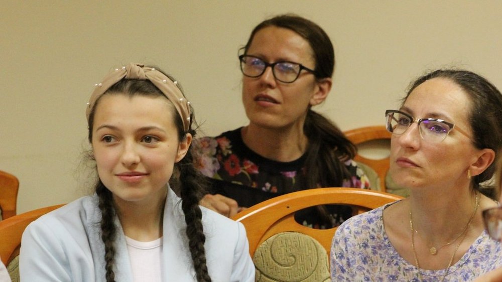 Trzy kobiety siedzące obok siebie. Z lewej strony to młoda dziewczyna z dwoma warkoczami, ubrana w jasną bluzę i opaskę na głowie. Obok niej siedzi kobieta w okularach i kolorowej bluzce w kwiaty. Po prawej stronie jest kolejna kobieta w okularach, ubrana w jasną bluzkę w kwiatowy wzór. Wszystkie trzy kobiety patrzą w jednym kierunku, prawdopodobnie na osobę mówiącą lub prezentację.