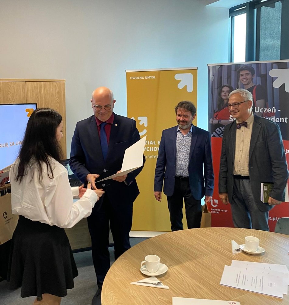 Cztery osoby w sali konferencyjnej. Trzech mężczyzn w garniturach stoi z prawej strony, a jeden z nich wręcza certyfikat młodej kobiecie po lewej stronie, która jest ubrana w białą bluzkę i czarną spódnicę. W tle znajdują się dwa roll-upy reklamowe: jeden żółty z logo Wydziału Nauk o Wychowaniu, drugi czerwony z logo programu Zdolny uczeń-świetny student. 