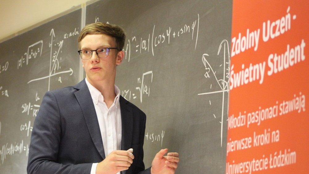 Młody mężczyzna stojący przed tablicą, na której zapisane są równania matematyczne i wykresy. Mężczyzna nosi okulary i jest ubrany w elegancką marynarkę oraz białą koszulę. Wygląda na skupionego i zaangażowanego, trzyma kredę w dłoni i patrzy w bok, jakby tłumaczył coś niewidocznej grupie osób. Po prawej stronie zdjęcia widoczny jest czerwony roll-up z napisem "Zdolny Uczeń - Świetny Student".