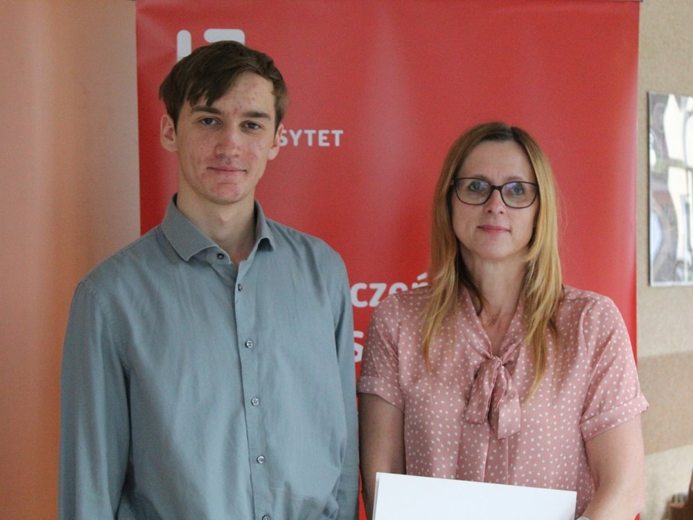 Dwie osoby stojące przed czerwonym roll-upem z napisem "Uniwersytet Łódzki". Po lewej stronie młody mężczyzna ubrany w jasnoniebieską koszulę, patrzący prosto w obiektyw z delikatnym uśmiechem. Po prawej stronie stoi kobieta w średnim wieku, ubrana w różową bluzkę z kokardą, nosząca okulary. Kobieta trzyma dokument.