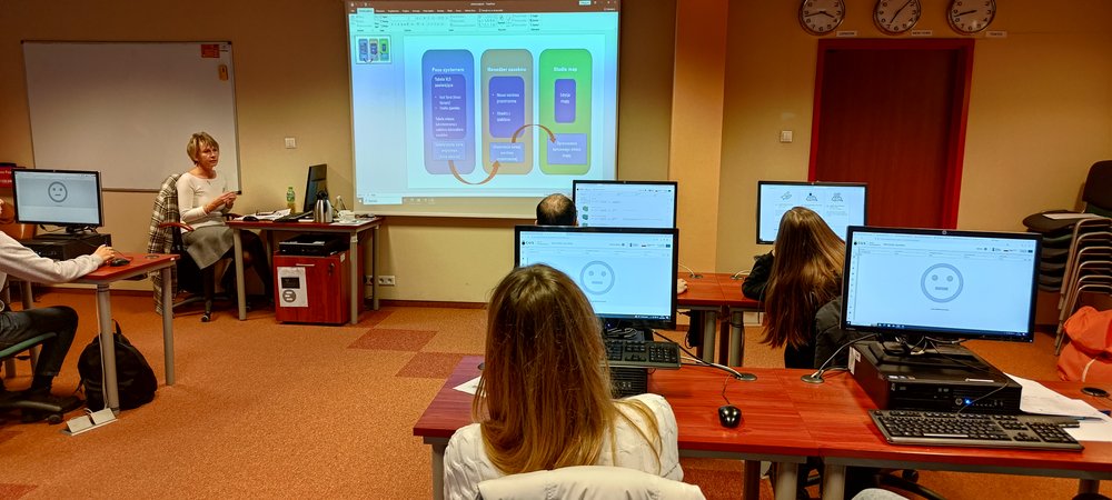 Prowadząca siedzi przy biurku z komputerem i omawia wyświetloną na dużym ekranie prezentację o Portalu Geostatystycznym