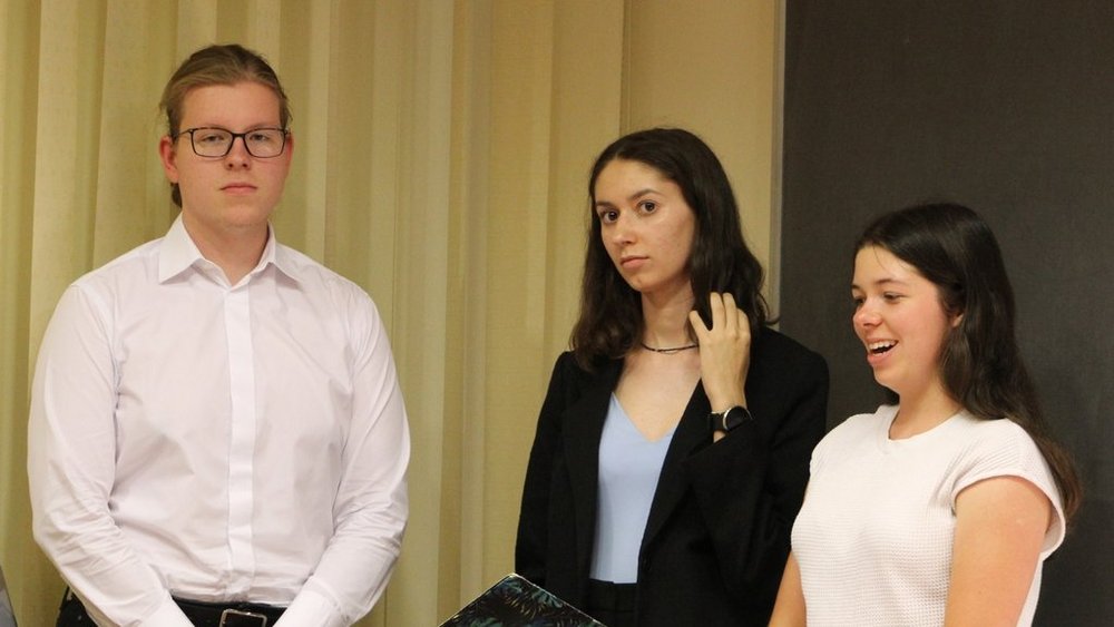 Trzy młode osoby stojące obok siebie. Po lewej stronie stoi chłopak w białej koszuli i okularach, z poważnym wyrazem twarzy. W środku stoi dziewczyna w czarnym żakiecie i jasnoniebieskiej bluzce, poprawiająca włosy, patrząc w bok. Po prawej stronie jest dziewczyna w białej koszulce, uśmiechająca się szeroko.  Tło zdjęcia stanowią jasne zasłony lub żaluzje oraz fragment ciemnej tablicy.