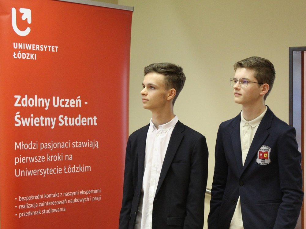 Dwaj młodzi mężczyźni stojący obok siebie. Są elegancko ubrani w marynarki i białe koszule. Mężczyźni patrzą w bok, w tym samym kierunku. W tle widoczny jest czerwony roll-up z napisem "Uniwersytet Łódzki" oraz napisem "Zdolny Uczeń - Świetny Student".