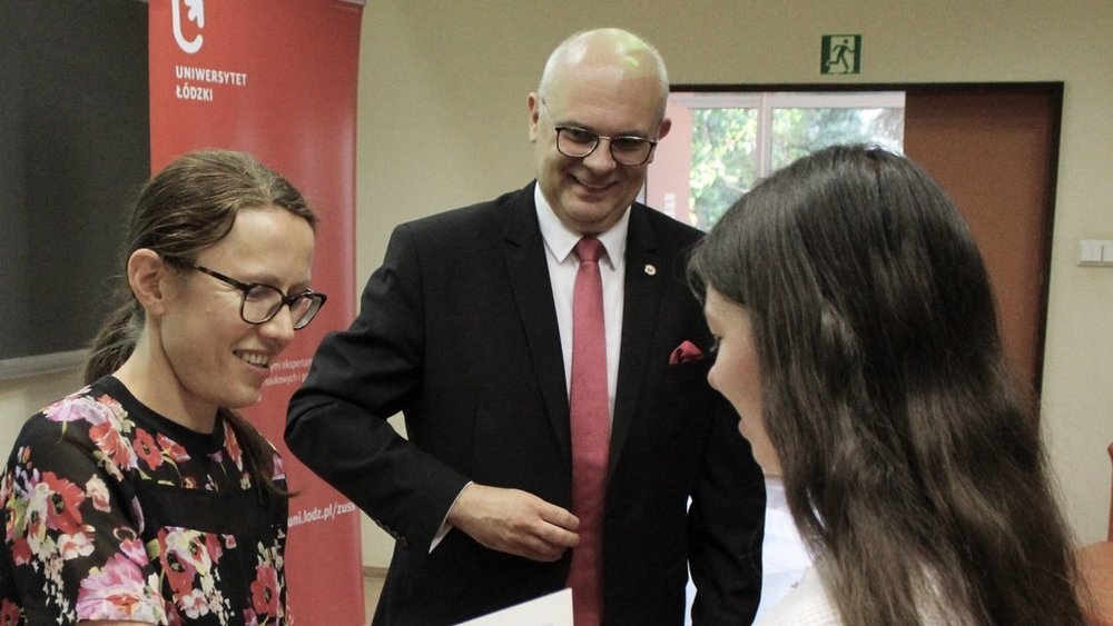 Na zdjęciu są trzy osoby. Po lewej stronie stoi kobieta w okularach i kolorowej bluzce w kwiaty, uśmiechająca się serdecznie. Obok niej, w centrum kadru, stoi mężczyzna w eleganckim garniturze z czerwoną krawatą i białą koszulą, również uśmiechnięty. Po prawej stronie znajduje się młoda kobieta z długimi, ciemnymi włosami, patrząca na certyfikat, który trzyma w rękach. W tle widoczny jest czerwony roll-up z logo "Uniwersytet Łódzki". 