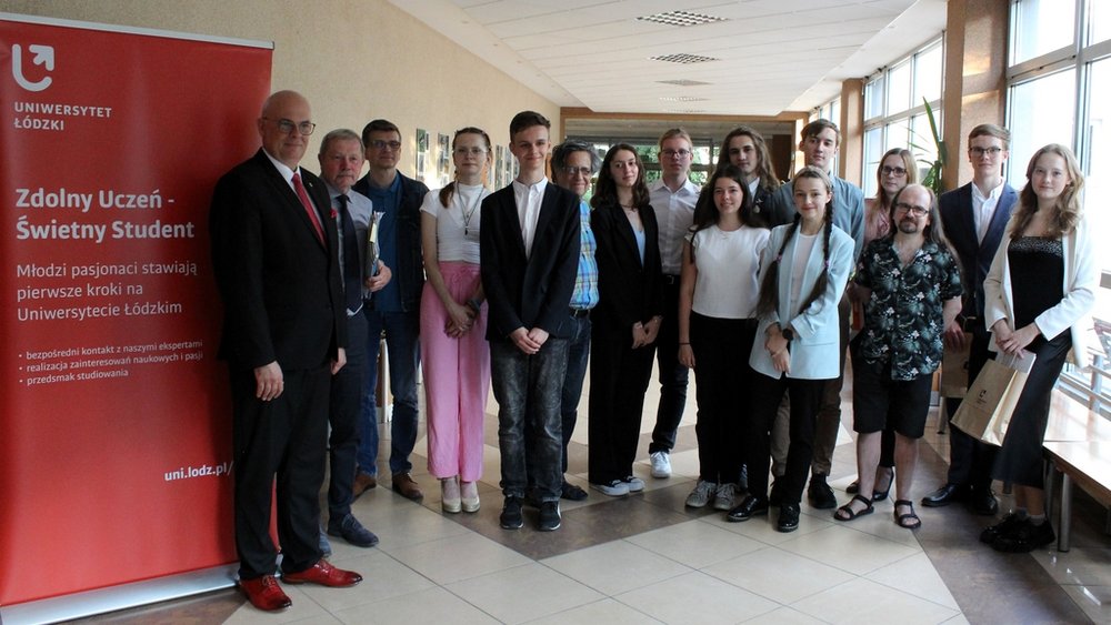 Grupa młodych i starszych osób  stojących w korytarzu budynku. Wszyscy są elegancko ubrani, niektórzy z nich trzymają dokumenty. Po lewej stronie widoczny jest czerwony roll-up z napisem "Zdolny Uczeń - Świetny Student" . W tle widać okna, przez które wpada naturalne światło.