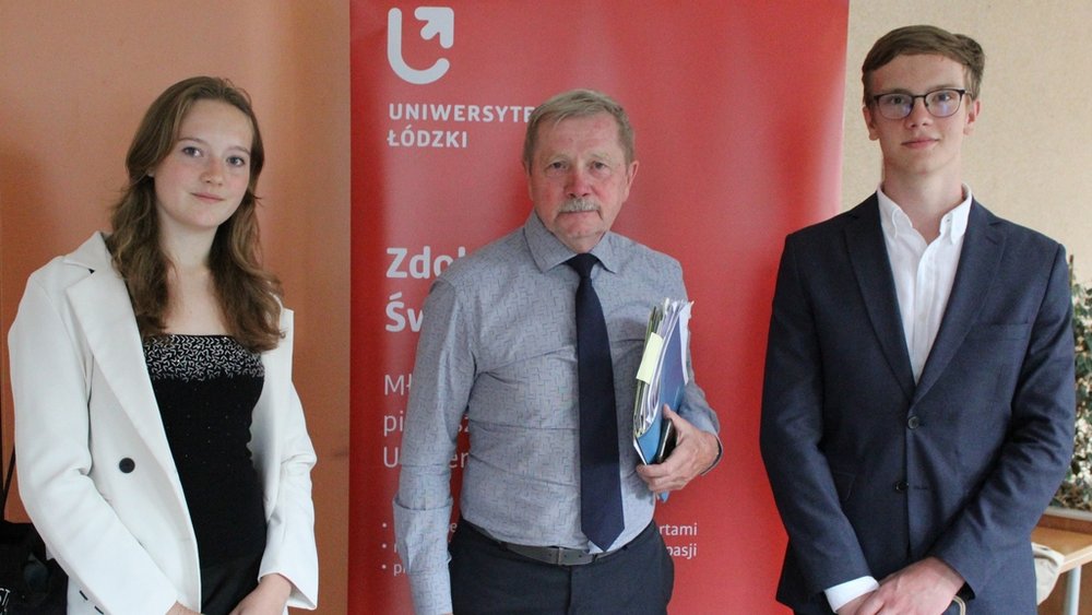 Trzy osoby stojące przed czerwonym roll-upem z napisem "Uniwersytet Łódzki". Po lewej stronie stoi młoda kobieta z długimi, jasnobrązowymi włosami, ubrana w jasną marynarkę i ciemną bluzkę, uśmiechająca się delikatnie do kamery. W środku znajduje się starszy mężczyzna z wąsami, ubrany w niebieską koszulę i ciemne spodnie, trzymający w rękach dokumenty lub książki. Po prawej stronie stoi młody mężczyzna w okularach, ubrany w elegancką marynarkę i białą koszulę, również uśmiechnięty.