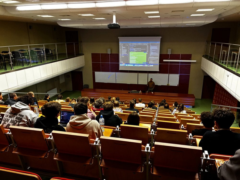 Na zdjęciu widoczna jest duża sala wykładowa oglądana z tylnej części audytorium. W rzędach ustawionych schodkowo siedzi kilkadziesiąt osób – głównie studenci – skierowanych w stronę prowadzącego.  Na dole sali, przy biurku, stoi wykładowca, który prowadzi prezentację. Nad nim znajduje się duży ekran projekcyjny, na którym wyświetlony jest kod komputerowy oraz interfejs programu, co wskazuje na tematykę informatyczną lub programistyczną.  Przed prowadzącym znajduje się długie biurko, a za nim tablice do pisania. Sala ma nowoczesny charakter – drewniane ławki, jasne oświetlenie oraz balkonowe poziomy po bokach, na których również ustawione są miejsca siedzące.  Studenci siedzą blisko siebie, część z nich ma otwarte laptopy lub notatki. Wszyscy zwróceni są w stronę wykładowcy, co sugeruje aktywne uczestnictwo w zajęciach. Atmosfera przypomina duży wykład akademicki lub prezentację specjalistyczną.