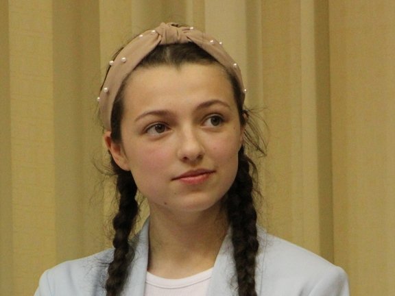 Młoda dziewczyna z długimi, ciemnymi włosami zaplecionymi w dwa warkocze. Ma na sobie jasną bluzę i białą koszulkę, a na głowie nosi jasną opaskę. Jej twarz wyraża delikatny uśmiech i zainteresowanie. 