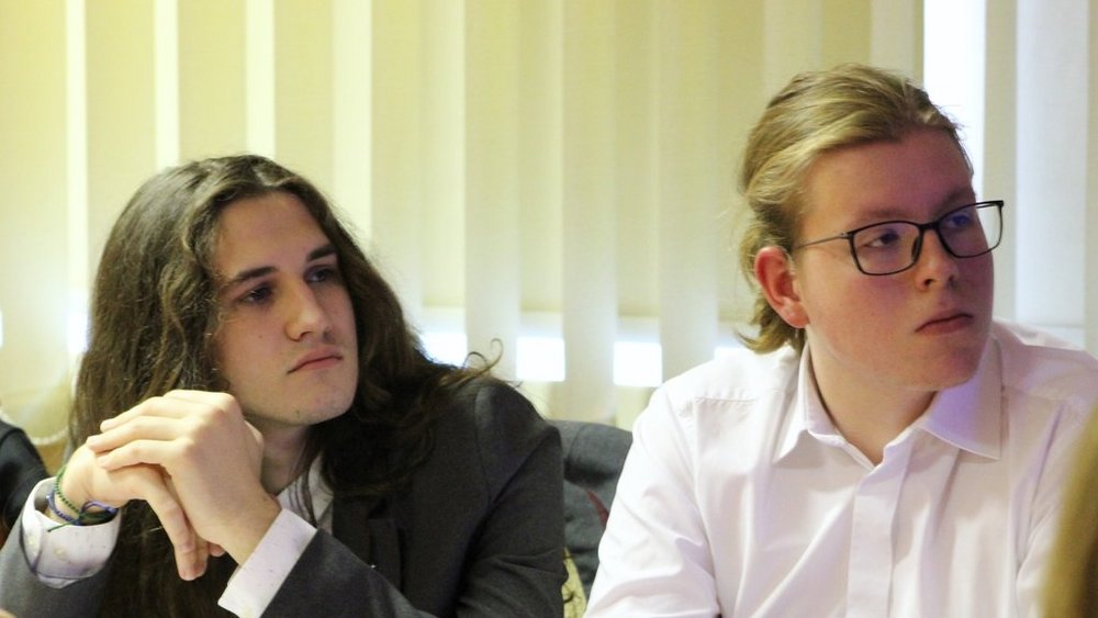 Dwaj młodzi mężczyźni siedzący obok siebie. Pierwszy z nich, po lewej stronie, ma długie, ciemne włosy i jest ubrany w garnitur. Opiera dłonie o stół i patrzy z zainteresowaniem w jednym kierunku. Drugi mężczyzna, po prawej stronie, ma krótkie, jasne włosy spięte w kucyk i nosi okulary. Jest ubrany w białą koszulę i również patrzy w tym samym kierunku. W tle widać jasne żaluzje, które przepuszczają naturalne światło.