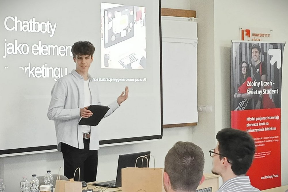 Na zdjęciu znajduje się młody mężczyzna  przed ekranem projekcyjnym, na którym wyświetlany jest slajd z napisem "Chatboty jako element marketingu". Prezenter trzyma tablet w jednej ręce i wskazuje na ekran drugą ręką. W tle, obok ekranu, znajduje się baner z napisem „Zdolny uczeń-świetny student”.