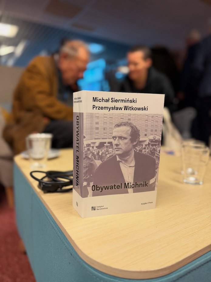 Zdjęcie ze spotkania, okładka nas stole, w tle Adam Michnik