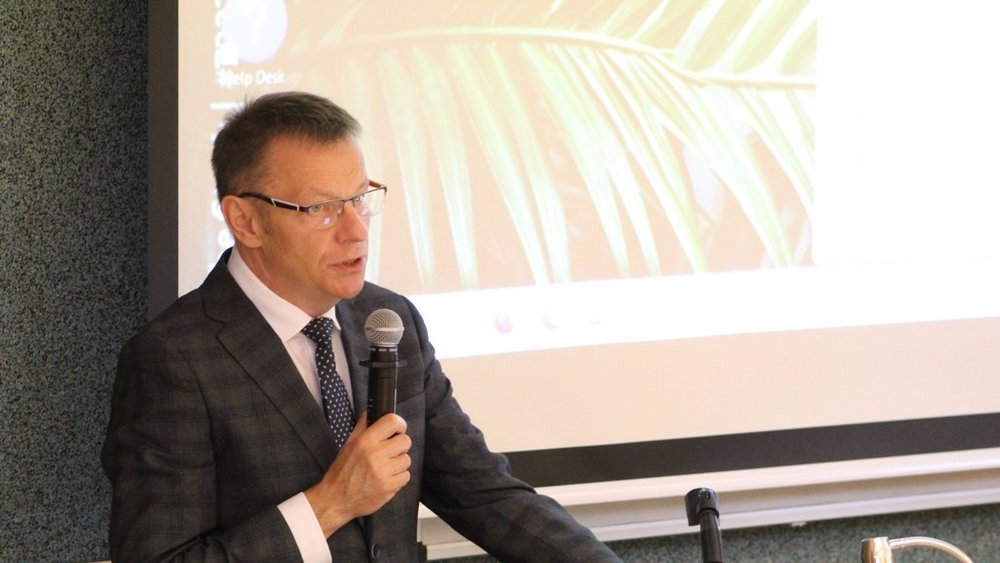 Mężczyzna w średnim wieku, elegancko ubrany w garnitur, koszulę i krawat. Mężczyzna trzyma mikrofon i wydaje się być w trakcie przemówienia. Patrzy przed siebie. W tle widoczny jest ekran projekcyjny, na którym wyświetlany jest slajd z motywem roślinnym.