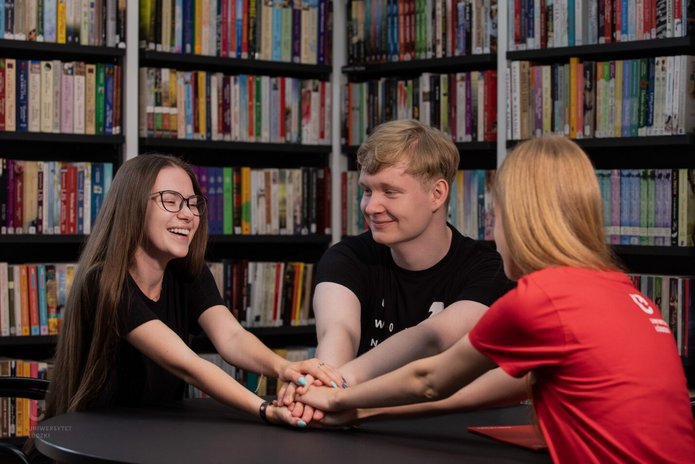 Kolorowe zdjęcie przedstawiające troje uśmiechniętych studentów siedzących na tle bibliotecznego księgozbioru. Na zdjęciu znajdują się dwie kobiety i jeden mężczyzna. Cała trójka trzyma się za ręce w znaku gotowości do wspólnego podjęcia wyzwania. Zdjęcie ma ciepły i jednocześnie motywacyjny charakter