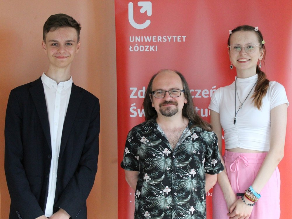 Po lewej stronie stoi młody mężczyzna ubrany w elegancką marynarkę i białą koszulę, z krótkimi, ciemnymi włosami. Po prawej stronie jest młoda dziewczyna z długimi, jasnymi włosami spiętymi w wysoki kucyk, ubrana w białą bluzkę i różową spódnicę. W środku stoi mężczyzna w średnim wieku, z długimi włosami i okularami, ubrany w koszulę z ciemnym, wzorzystym nadrukiem. Wszyscy wyglądają na zadowolonych.