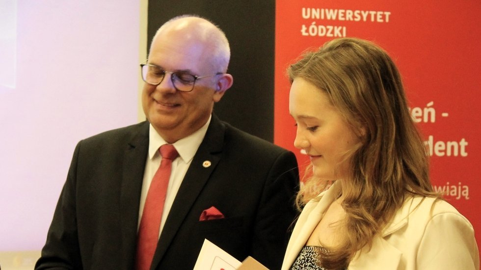 Na zdjęciu znajduje się mężczyzna i młoda kobieta. Mężczyzna jest elegancko ubrany w ciemny garnitur, białą koszulę i czerwoną krawatkę. Ma siwe włosy, nosi okulary i uśmiecha się serdecznie. Obok niego stoi młoda kobieta z długimi, jasnymi włosami, ubrana w jasną marynarkę. Trzyma dokument lub certyfikat i również uśmiecha się, patrząc w dół na trzymany przedmiot. W tle widać czerwony roll-up z napisem "Uniwersytet Łódzki" oraz częściowy napis "Zdolny Uczeń - Świetny Student".