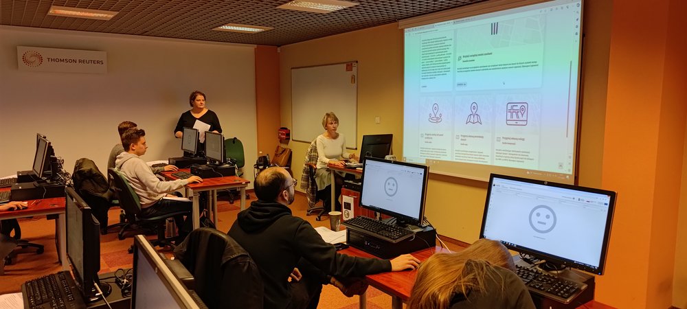 Dwie Prowadzące szkolenie o Portalu Geostatystycznym pokazują na dużym ekranie funkcjonalności Portalu; uczestnicy szkolenia siedzą przy komputerach i słuchają