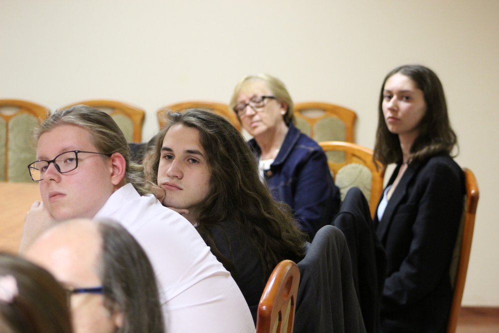 Grupa młodych osób siedzących przy stole, prawdopodobnie w sali konferencyjnej lub klasie. Pierwszy plan zajmują trzej młodzi ludzie: chłopak z jasnymi, spiętymi włosami i okularami, obok niego chłopak z długimi, ciemnymi włosami oraz dziewczyna z ciemnymi włosami, ubrana w ciemny strój. W tle siedzi starsza kobieta w okularach, również skupiona na tym, co się dzieje. Wszyscy patrzą w jednym kierunku, co sugeruje, że słuchają prezentacji.