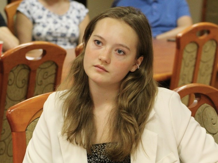  Młoda kobieta siedząca na krześle w Sali.  Ma długie, jasnobrązowe włosy i jest ubrana w jasną marynarkę oraz ciemną bluzkę. Wygląda na zamyśloną i skupioną, patrzy w dal, najprawdopodobniej słuchając kogoś, kto przemawia. W tle widać inne osoby siedzące przy stołach, a także drewniane krzesła z tapicerowanymi oparciami. 