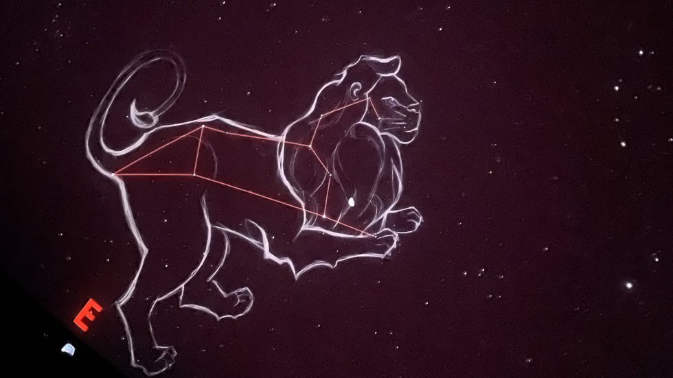 Na ciemnym, gwiaździstym tle widoczna jest ilustracja konstelacji Lwa. Gwiazdy połączone są cienkimi, czerwonymi liniami tworzącymi charakterystyczny kształt, a całość otacza jasny, szkicowy kontur przedstawiający lwa w pozycji stojącej. Po lewej stronie znajduje się czerwona litera „E”, oznaczająca kierunek wschodni. Tło jest jednolicie ciemne, a rozproszone punkty światła przedstawiają gwiazdy.