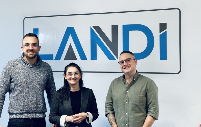 Dwóch pracowników firmy Landi oraz pracowniczka EkSocu stoją na tle dużego logotypu firmy. Od lewej: mgr Jan Fudali, dr Joanna Górniak i mgr Mateusz Piątkowski