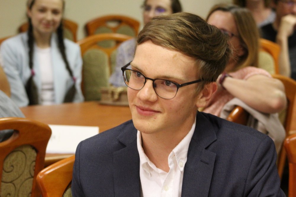 Młody mężczyzna siedzący przy stole. Ma krótkie, ciemne włosy i nosi okulary. Jest ubrany w elegancką marynarkę i białą koszulę. Mężczyzna uśmiecha się lekko i patrzy w stronę rozmówcy. W tle widać inne osoby, które również siedzą przy stołach i słuchają.