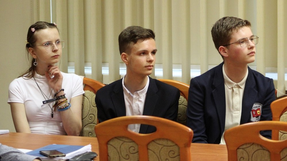 Trzy młode osoby siedzące przy stole. Po lewej stronie siedzi dziewczyna z długimi, jasnymi włosami, ubrana w białą koszulkę. Trzyma rękę przy brodzie, sprawiając wrażenie zamyślonej. Obok niej siedzi dwóch chłopców, obaj elegancko ubrani w marynarki i białe koszule. Wszyscy patrzą w jednym kierunku, prawdopodobnie na osobę mówiącą lub prezentację. W tle widać żaluzje na oknach.