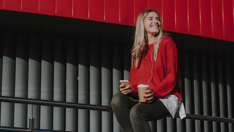 Na zdjęciu widoczna jest młoda kobieta siedząca na metalowej barierce. Ma na sobie czerwony sweter, ciemne spodnie i białą koszulę wystającą spod swetra. W jednej dłoni trzyma smartfon, w drugiej papierowy kubek z kawą; w uszach ma słuchawki. Kobieta uśmiecha się i patrzy w bok. Tło stanowi nowoczesna architektura z pionowymi, ciemnymi panelami oraz intensywnie czerwonym elementem konstrukcyjnym u góry kadru.