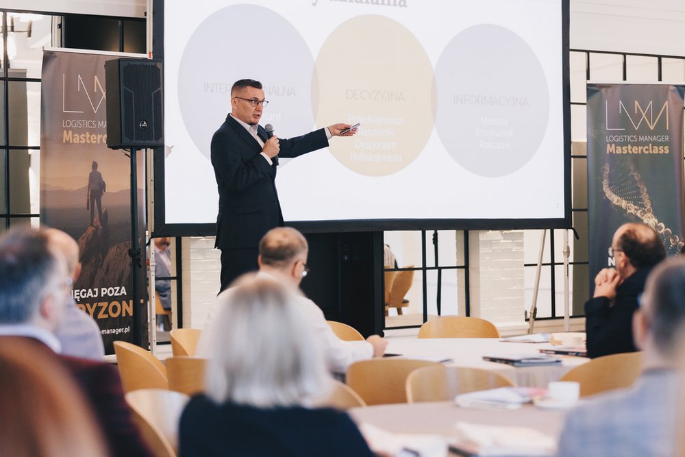 mężczyzna przemawiający przed publicznością, a za nim ekran z prezentacją