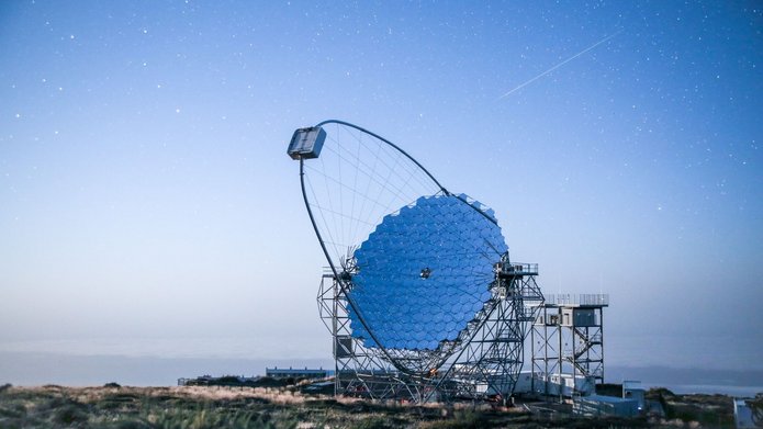 Pojedynczy teleskop astronomiczny z dużym, segmentowym zwierciadłem i stalową konstrukcją wsporczą na górskim szczycie. Nad teleskopem rozciąga się błękitne, rozgwieżdżone niebo, a w tle widoczna jest warstwa chmur.