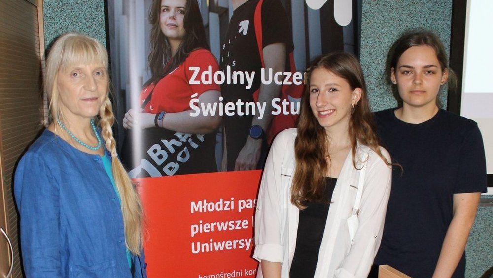 Na zdjęciu znajdują się trzy kobiety stojące obok siebie przed plakatem z napisem "Zdolny Uczeń - Świetny Student". Po lewej stronie stoi starsza kobieta z jasnymi włosami związanymi w warkocz, ubrana w niebieską bluzkę. W środku stoi młoda dziewczyna z długimi, brązowymi włosami, ubrana w białą koszulę a po prawej stronie jest młoda kobieta o ciemnych włosach, ubrana w czarną bluzkę, która ma poważny wyraz twarzy.