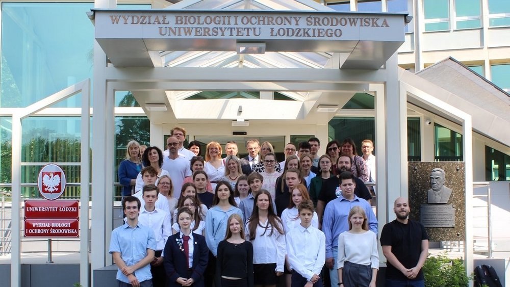 Duża grupa osób stojących przed budynkiem oznaczonym napisem "Wydział Biologii i Ochrony Środowiska Uniwersytetu Łódzkiego". Grupa składa się z młodych ludzi i dorosłych. Wszyscy są elegancko ubrani, niektórzy w białe koszule, inni w ciemne marynarki.W tle widać przeszklone drzwi budynku oraz tabliczkę z godłem Polski po lewej stronie.