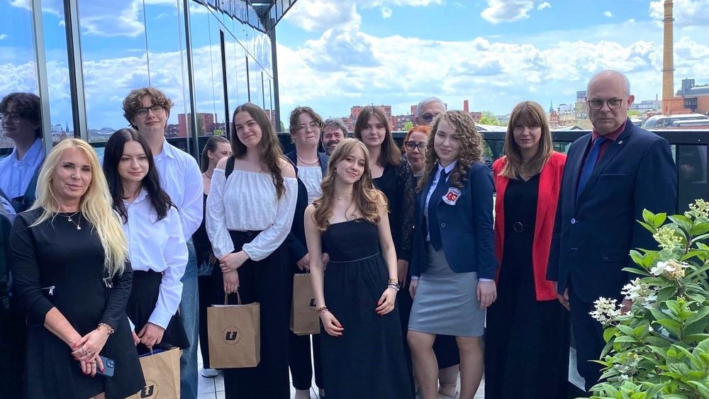 Grupa osób stojących na zewnątrz, na tle budynku z przeszklonymi ścianami. W skład grupy wchodzi zarówno młodzież, jak i dorośli. Wszyscy są elegancko ubrani. Wiele osób trzyma torby z logo Uniwersytetu Łódzkiego. W tle widać niebo z chmurami oraz fragmenty miejskiego krajobrazu z budynkami. 