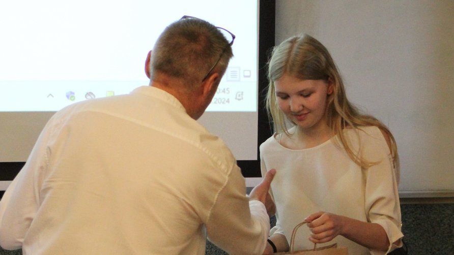 Na zdjęciu widoczna jest młoda kobieta o jasnych, długich włosach, ubrana w jasną bluzkę. Odbiera coś od starszego mężczyzny, który jest zwrócony do niej plecami i ubrany w białą koszulę. Kobieta trzyma w rękach papierową torbę i uśmiecha się delikatnie, patrząc w dół. Tło stanowi ekran projekcyjny, na którym wyświetlony jest fragment prezentacji