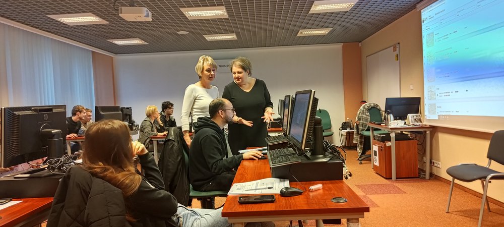 Dwie Prowadzące stoją przy jednym z uczestników wykonującym zlecone zadanie na komputerze i próbują mu wyjaśnić jak to zrobić