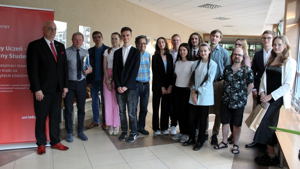 Grupa osób w młodszym i starszym wieku stojących w korytarzu budynku. Wszyscy są elegancko ubrani, niektórzy trzymają dokumenty lub torby. Po lewej stronie widoczny jest czerwony roll-up z napisem "Zdolny Uczeń - Świetny Student" oraz logo Uniwersytetu Łódzkiego. Obok roll-upu stoi mężczyzna w ciemnym garniturze z czerwoną koszulą i czerwonymi butami.