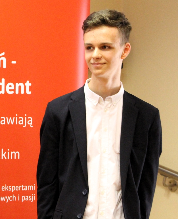 Młody mężczyzna stojący przed czerwonym roll-upem z napisem promującym program "Zdolny Uczeń - Świetny Student". Mężczyzna jest elegancko ubrany w białą koszulę i ciemną marynarkę. Ma krótkie, ciemne włosy i uśmiecha się delikatnie, patrząc w bok.